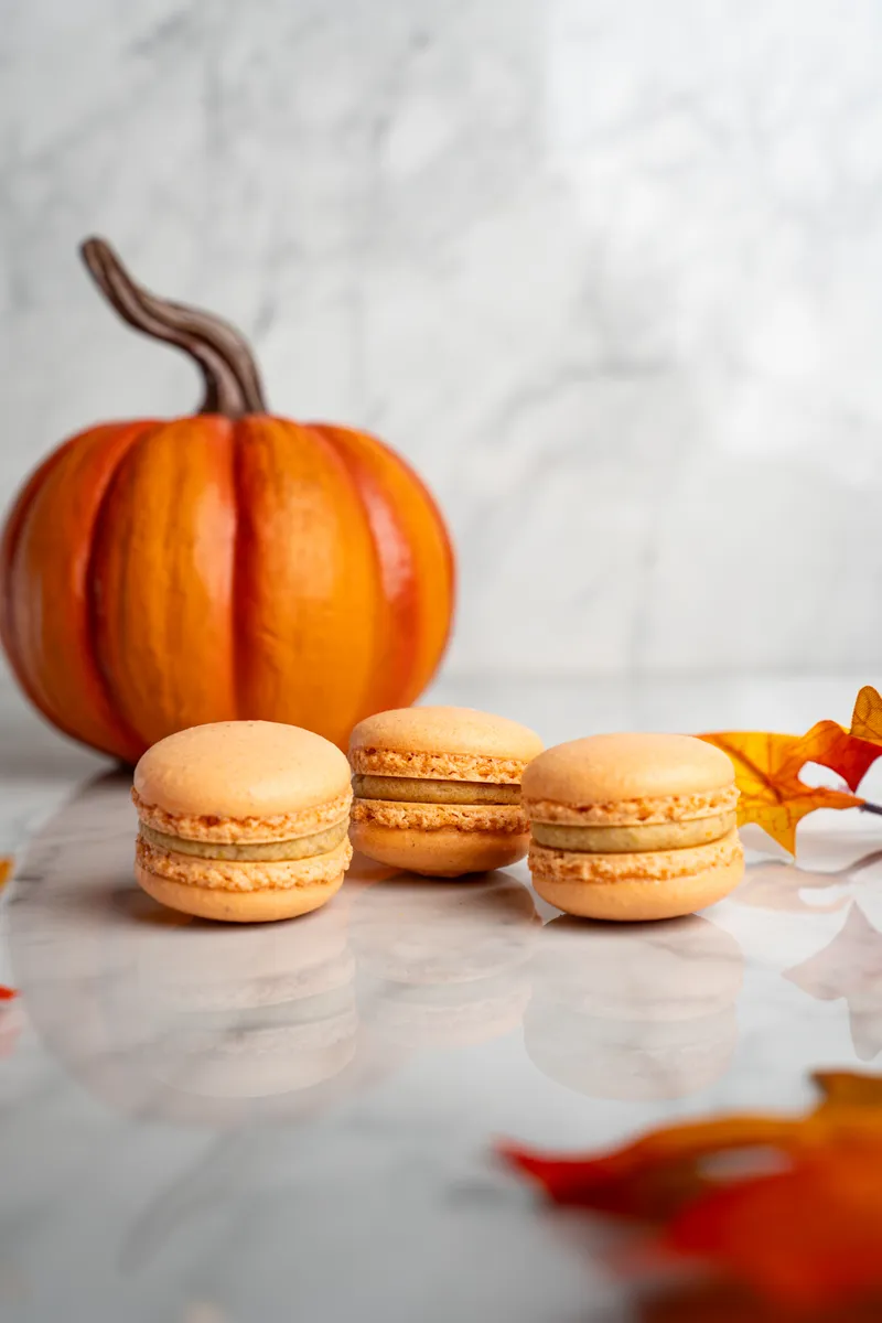 Orange pumpkin spice macarons on marble with a small pumpkin behind them